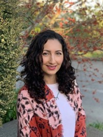 A woman with long black hair and red shawl smiling in an outdoor setting. The background includes autumn trees and foliage.