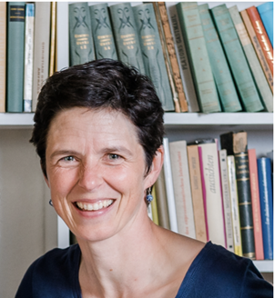 A woman with cropped hair, blue eyes and small blue earrings smiling in front of a bookshelf.