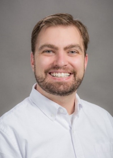 A man with cropped blond hair and facial hair in a white shirt, smiling in front of a gray background.