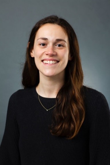 A woman with long brown hair in a black long sleeve and a small necklace smiling in front of a dark gray background. 