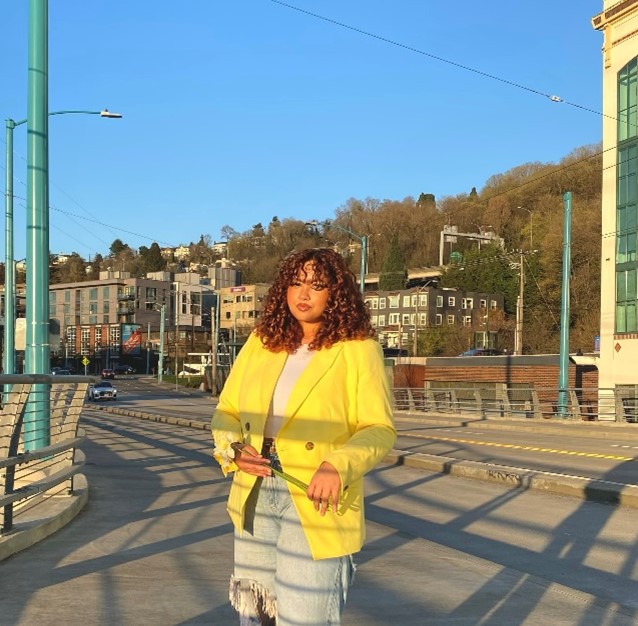 A woman with curly auburn bangs, a yellow blazer, and jeans stands on a sidewalk at sunset. 